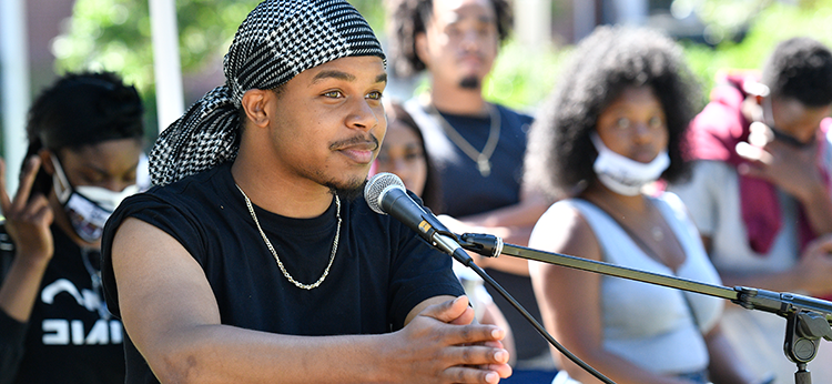 A young African American man gives a speech during an outdoor event at 爱妹社. Behind him, blurred, are several young African American male and female students.