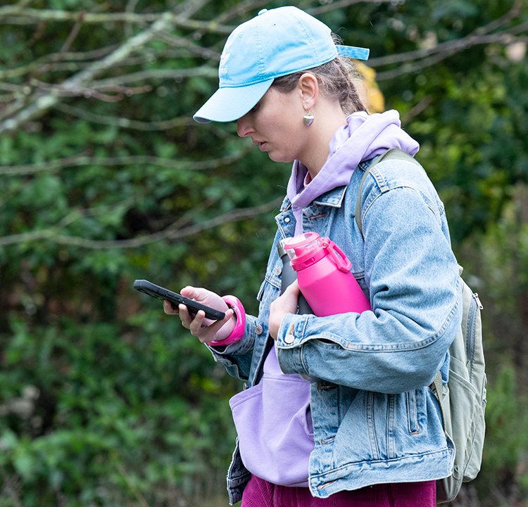A young blond woman is looking at her phone while walking on the Santa Rosa campus. She is wearing a denim cap and jacket and is holding a pink water bottle.